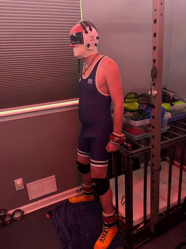 Me, wearing a purple wrestling singlet, yellow sneakers, and white headgear. I am blindfolded and gagged with tape, and my wrists and ankles are chained to a cage, holding me in place.