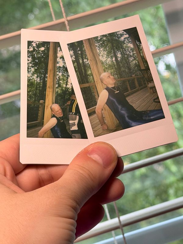 Two polaroids being held in someone's hand. The pictures depict me, sitting against a post with my hands cuffed behind me, I am wearing a blue amoresy suit and I am covered in piss