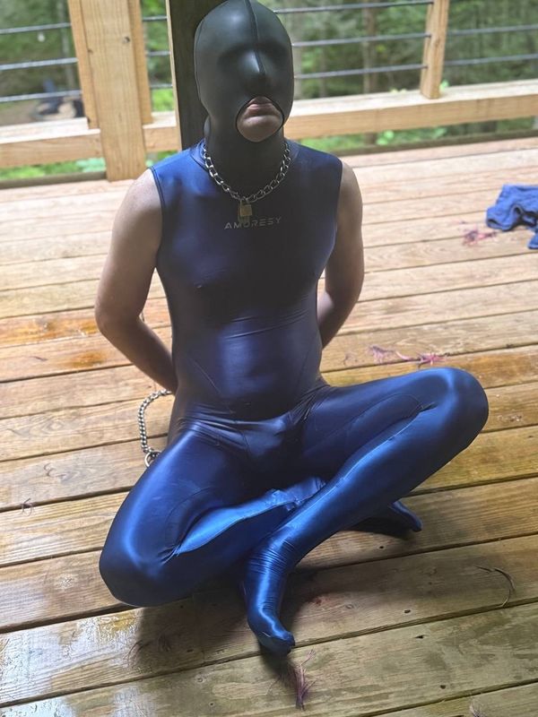 me, wearing a blue sleeveless amoresy swimsuit. I am sitting on a wooden deck, and my wrists are cuffed around a post behind my back. I am drenched in piss, and wearing a hood with just a mouth opening